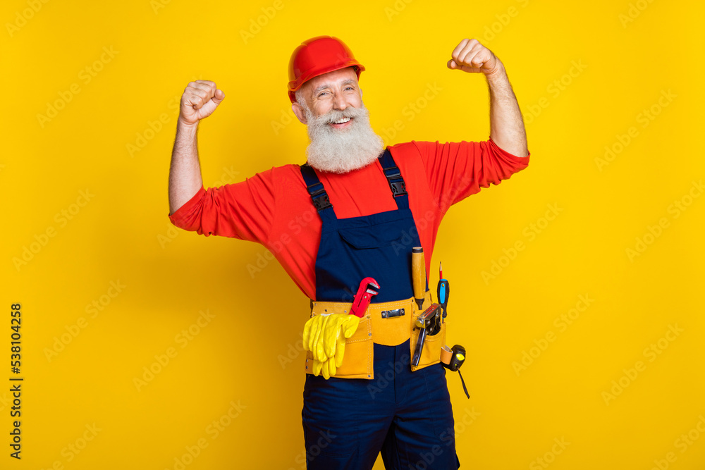Photo of cool cheerful age man wear overall uniform red hard hat showing biceps rising fists isolated yellow color background