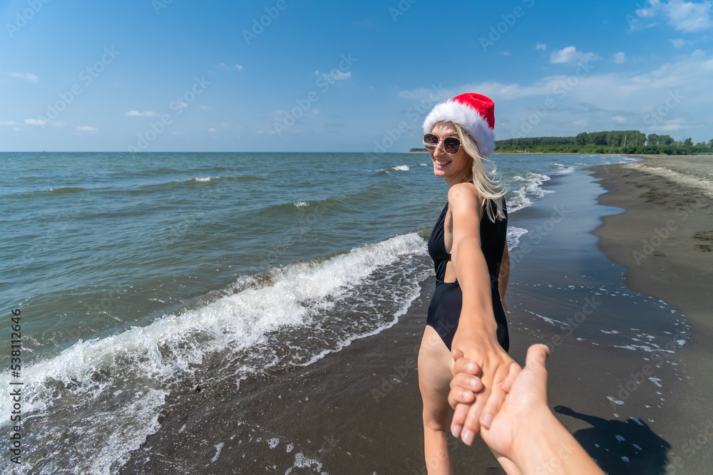 Young smiling slender blonde woman in a black swimsuit, sunglasses, Santa Claus hat holds a man's hand and walks along the seashore on a sunny day. Follow me Young smiling slender blonde woman in a black swimsuit, sunglasses, Santa Claus hat holds a man's hand and walks along the seashore on a sunny day. Follow me