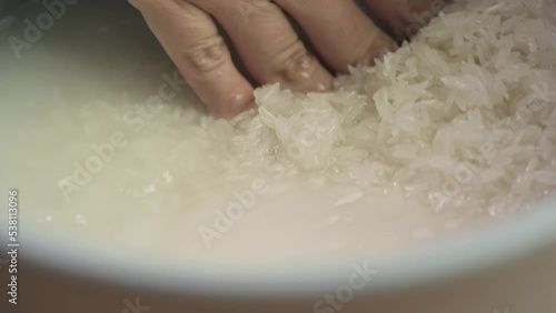 Close up of hand washing white Thai jasmine long grain rice with water in a rice cooker bowl in slow motion.