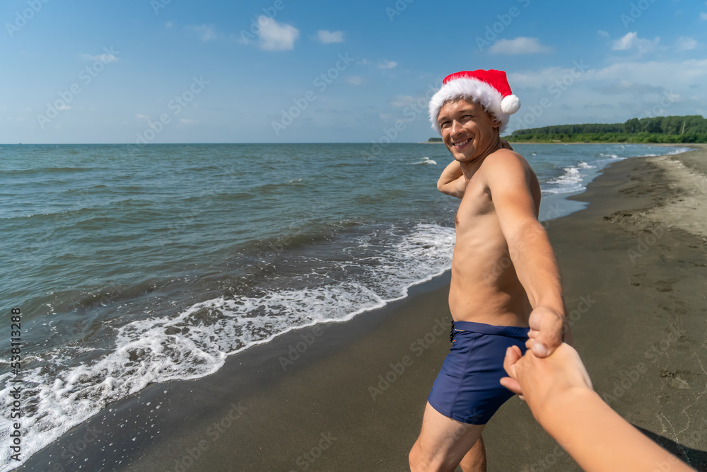 Happy smiling man in swimming trunks, Santa Claus hat holds a woman's hand and runs along the seashore on a sunny day. Come with me. Christmas holidays in a warm country Happy smiling man in swimming trunks, Santa Claus hat holds a woman's hand and runs along the seashore on a sunny day. Come with me. Christmas holidays in a warm country
