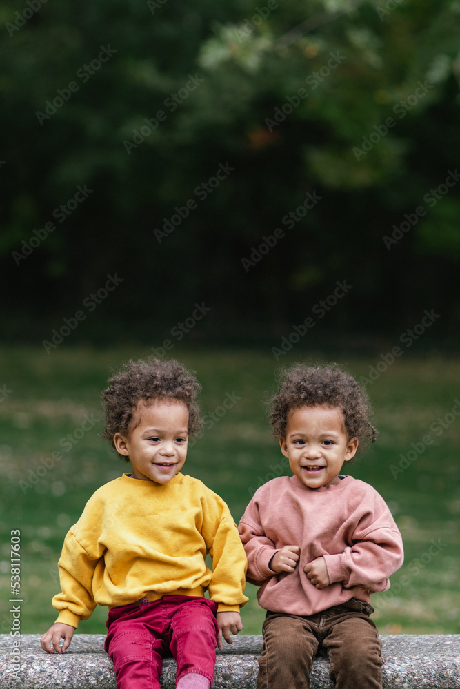 Black and Asian twins sitting on bench in park while smiling Black and Asian twins sitting on bench in park while smiling