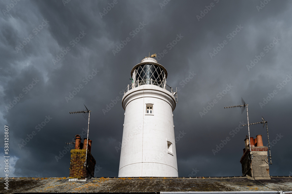 Dramatic sky seen against sun striking a lighthouse. Seen rising from nearby roofs. The lighthouse warns shipping of being run aground.