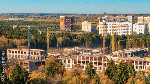 Construction site panorama. Construction business of real estate and residential buildings. Build in forest area in Troitsk city. Cranes against woods trees and city in autumn sunny day