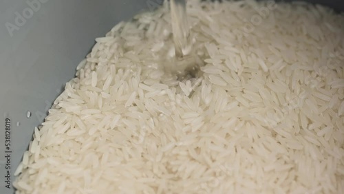 Pouring water into a rice cooker with raw white Thai jasmine long grain rice close up slow motion.