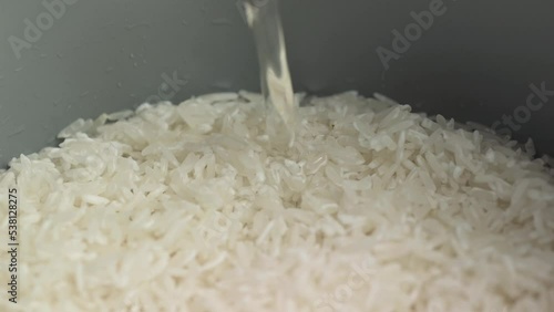 Pouring water into a rice cooker with raw white Thai jasmine long grain rice close up slow motion.
