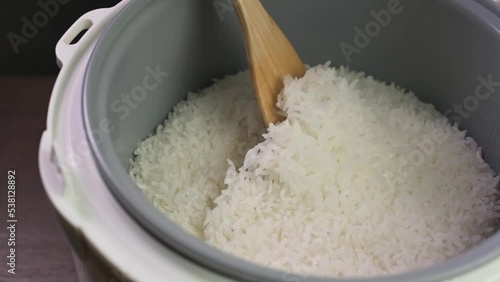 Wooden spoon stir cooked white Thai jasmine long grain rice in a rice cooker bowl close up slow motion.