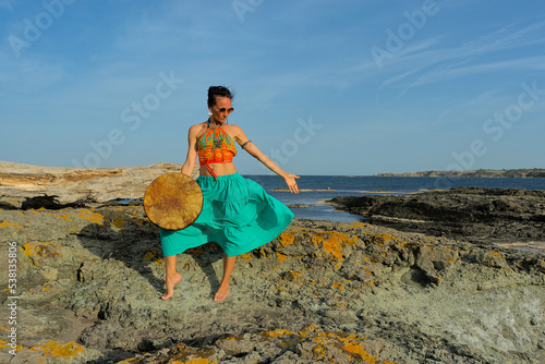 A woman in boho style clothes with a shamanic ritual tambourine on the rocks on the seashore on a clear sunny day. The concept of freedom of mind and body in nature