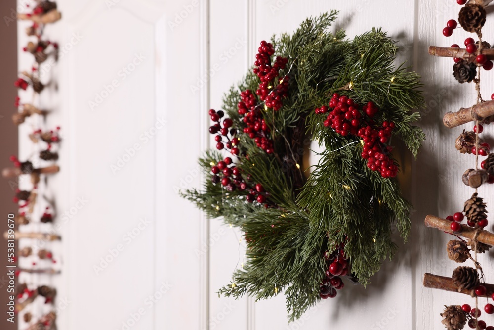 Beautiful Christmas wreath with red berries and fairy lights hanging on white door, space for text