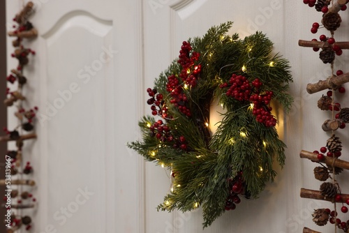 Beautiful Christmas wreath with red berries and fairy lights hanging on white door, space for text