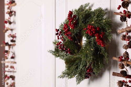 Beautiful Christmas wreath with red berries and fairy lights hanging on white door  space for text