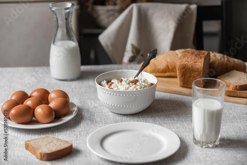 At home  in the kitchen  the table is covered with a linen tablecloth and there are natural and healthy products  milk and cottage cheese  bread and boiled chicken eggs on it
