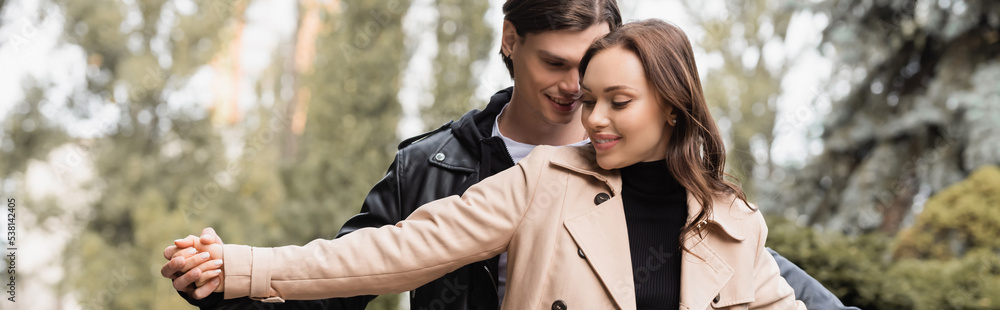 cheerful man holding hands with pleased girlfriend in beige trench coat in park, banner. cheerful man holding hands with pleased girlfriend in beige trench coat in park, banner.
