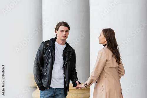 young man in stylish jacket holding hands with cheerful girlfriend in trench coat near white columns.