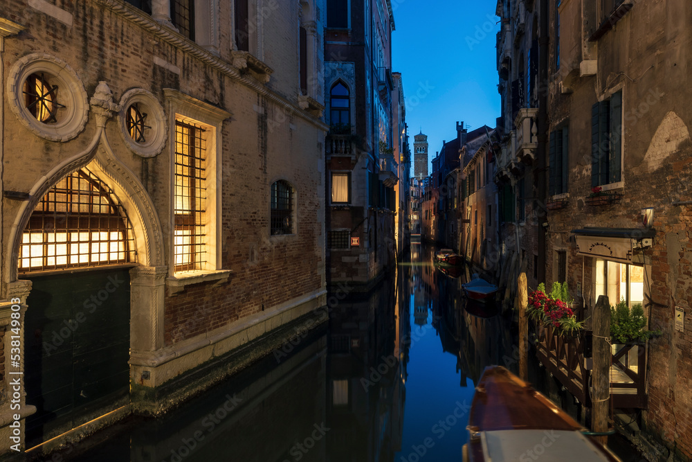 Venetian canal at night time, with the lights of the buildings reflecting in the calm waters