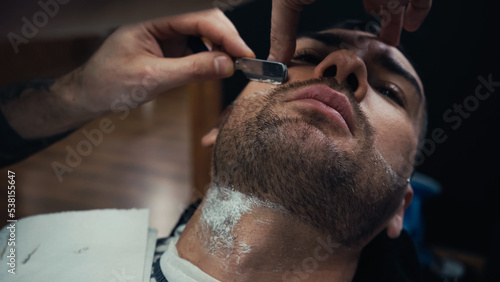 Barber shaving beard of man with razor and foam in salon.