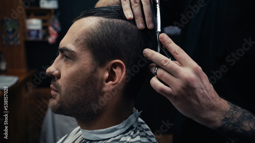 Hairdresser cutting hair of man in hairdressing cape in barbershop.