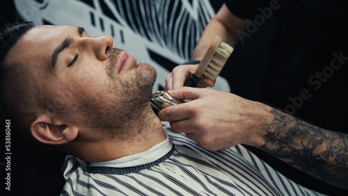 Tattooed barber holding brush and trimming beard of customer in salon.