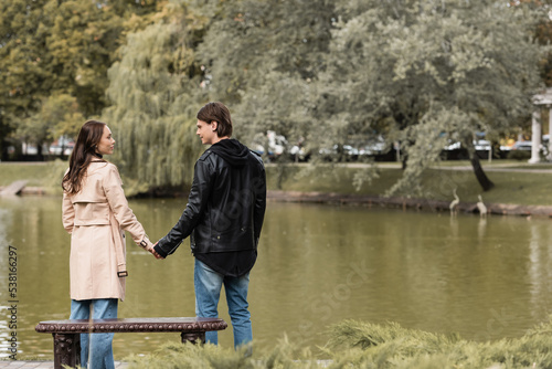 young couple in autumnal outfits holding hands while standing near lake in park.