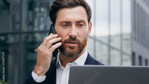 Multitasking man manager consultant talks on phone consulting client checks information on laptop businessman answering business call using smartphone makes order remote uses computer sitting outdoors