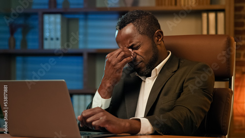Adult african american businessman working on laptop in office unhealthy tired man suffering from headache feels pain pressure fatigue from overwork suffer chronic migraine experiencing health problem
