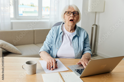 a shocked elderly lady is sitting at home in a cozy room at her desk with a laptop and looks up with her mouth wide open, thinking about solving work problems. The concept of working from home