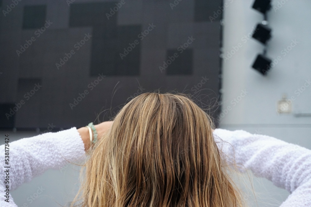Fototapeta premium Hinterkopf von junger Frau mit blonden langen Haaren, weißem Pullover und verschränkten Armen vor Kopf steht vor großer weißer Fassade mit schwarzer Leinwand auf Platz in Stadt am Morgen im Sommer