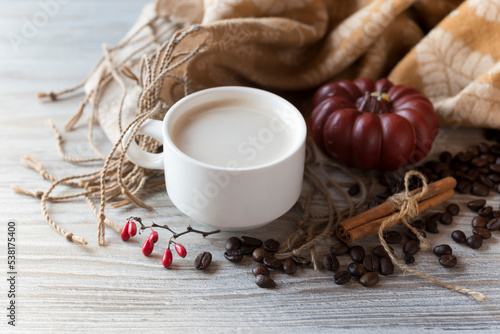 Cozy composition. Cup of coffee with cream, pumpkin candle, coffee beans, cinnamon sticks and fabric scarf on the light wooden background. Top view