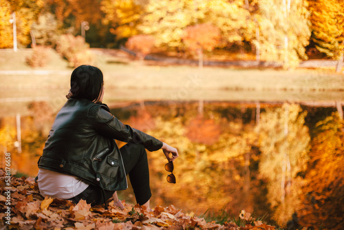 Young thoughtful stylish woman looking away while sitting by the lake in park during sunny weather in autumn. Orange trees reflected in the water