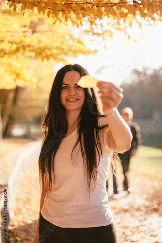 Happy smiling young woman holding yellow leaf in front of her face while standing in park against sunlight during sunny weather in autumn