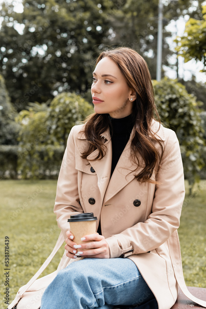 pretty young woman in beige trench coat sitting on bench with paper cup. pretty young woman in beige trench coat sitting on bench with paper cup.