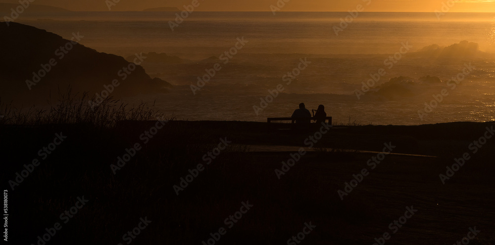 Silhouettes of two people sitting on a bench near the ocean at sunset
