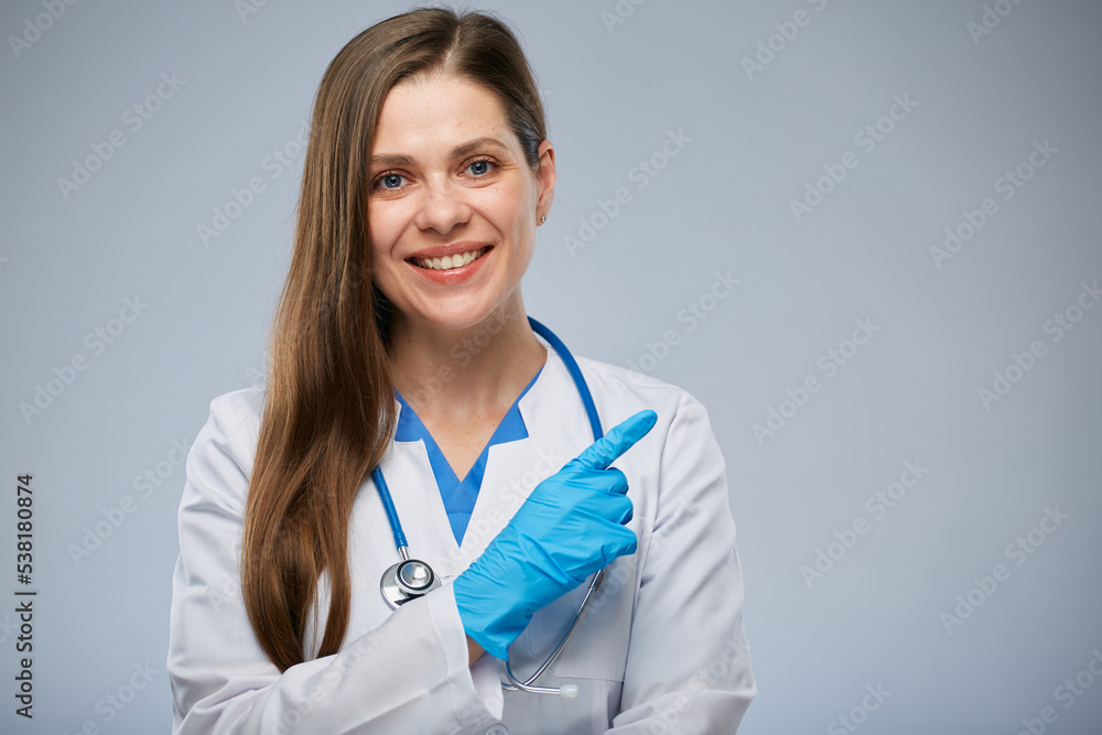Smiling doctor in medical white coat pointing finger at side. Isolated portrait of female medical worker