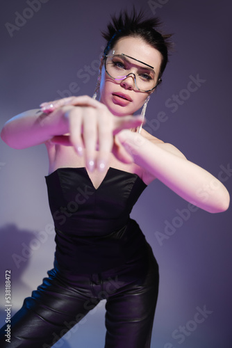 brunette woman in black corset and trendy sunglasses looking at camera and pointing with finger on purple background.