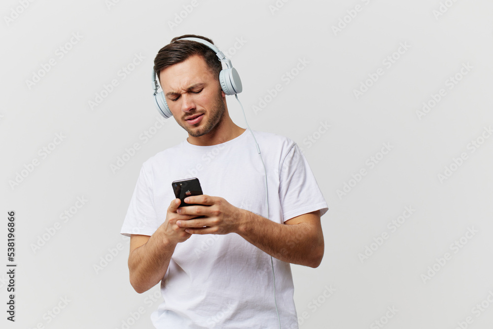 Happy concentrated tanned handsome man in basic t-shirt headphones listen playlist chatting with phone posing isolated on over white studio background. Copy space Banner Mockup. Music pause concept Happy concentrated tanned handsome man in basic t-shirt headphones listen playlist chatting with phone posing isolated on over white studio background. Copy space Banner Mockup. Music pause concept