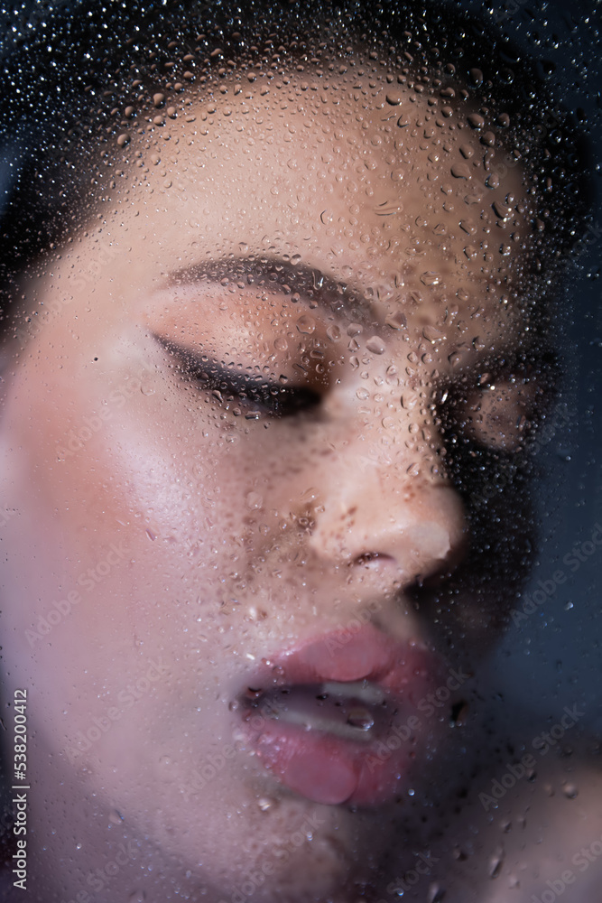 Blurred seductive woman standing near wet glass on grey background.