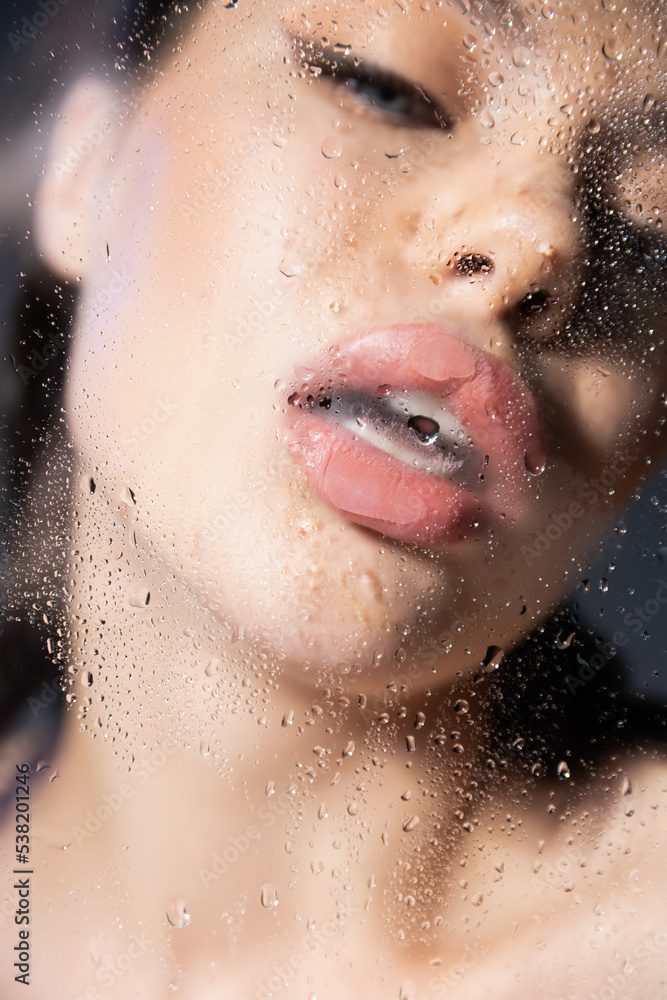 Blurred young woman with opened mouth near wet glass on grey background.