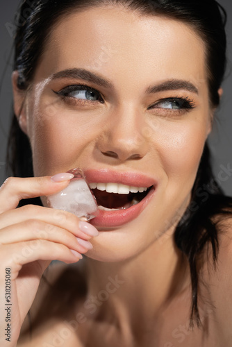 Cheerful brunette woman holding ice cube near lips isolated on grey.
