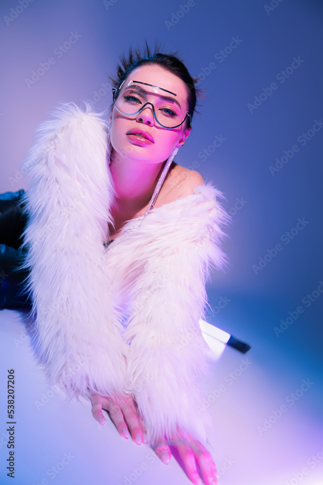 trendy woman in white faux fur jacket lying near fluorescent lamp and looking at camera on blue and purple background. trendy woman in white faux fur jacket lying near fluorescent lamp and looking at camera on blue and purple background.