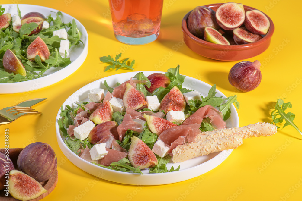 Autumn figs salad with arugula, feta, raw ham in white plate on yellow background served with rose wine and grissini. Flexitarian diet, paleo diet fall salad, close up Autumn figs salad with arugula, feta, raw ham in white plate on yellow background served with rose wine and grissini. Flexitarian diet, paleo diet fall salad, close up