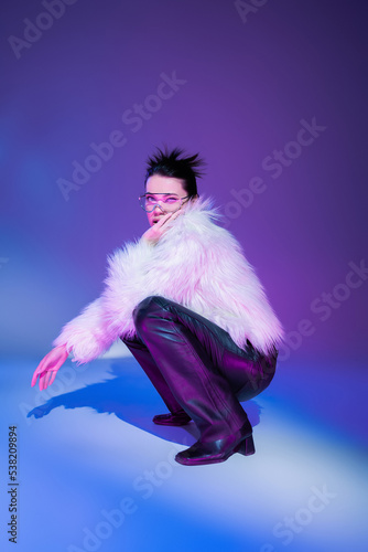 full length of brunette model in black leather pants and white furry jacket looking at camera on purple background.