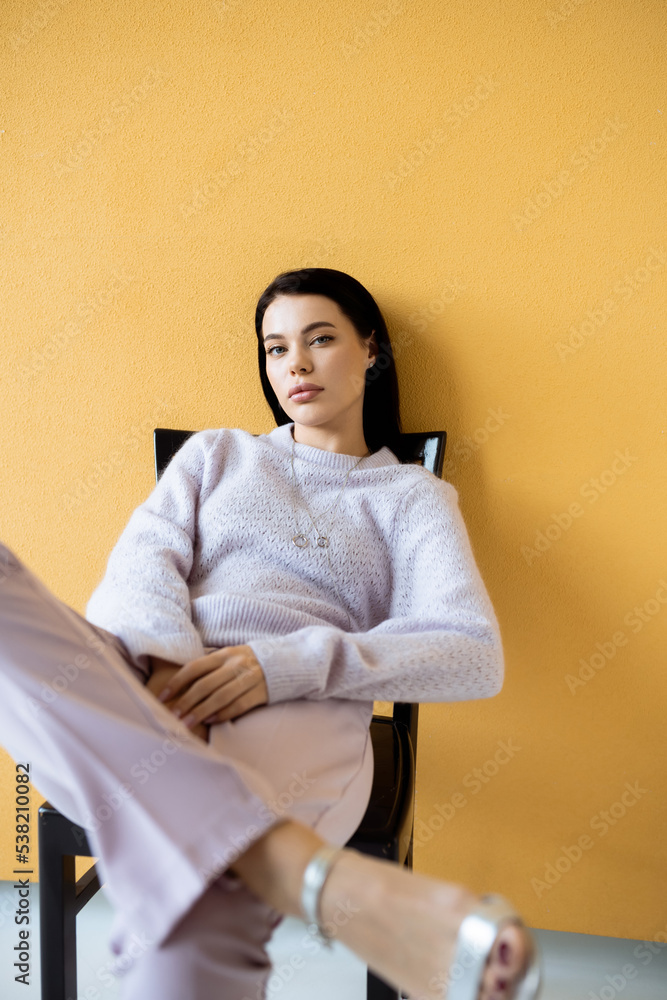 Obraz premium trendy woman in cozy sweater looking at camera while sitting on yellow background.