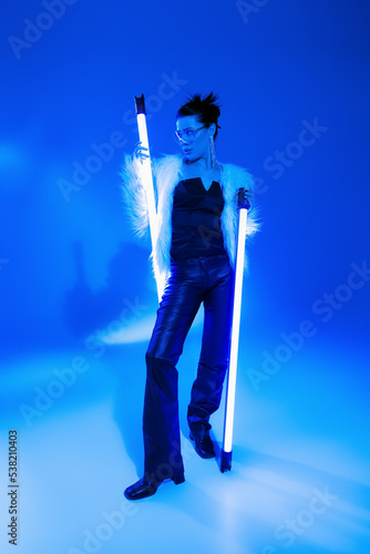 Stylish woman in furry jacket and sunglasses holding lamps on blue background.
