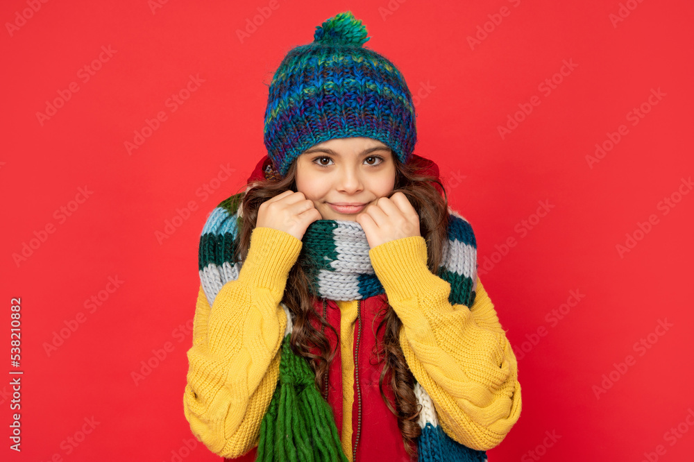 smiling child in knitted winter hat and scarf on red background, season fashion