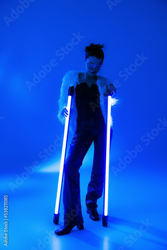 Full length of trendy woman in faux fur jacket holding lamps on blue background.