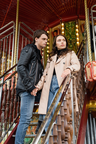 low angle view of stylish couple holding hands while standing on carousel in amusement park.