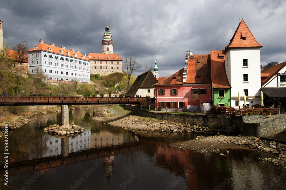 Cesky Krumlov, Czech Republic - April 16th 2022: View over Moldova to castel and tower with foot bridge, dark grey thunderstorm clouds 
