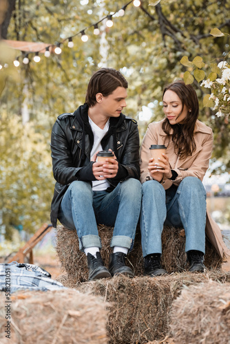 young woman and man in stylish outfits holding paper cups and sitting in autumnal park.