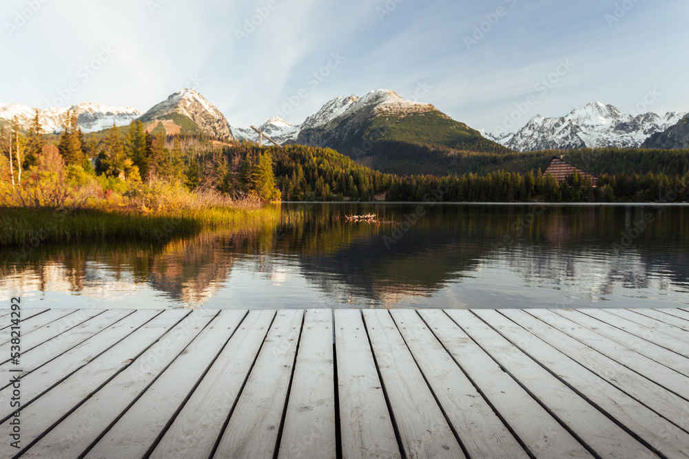 Mole (pier) on the lake in autumn scenery with beautiful and snowy mountains on background. Strbske pleso in High Tatras in fall with wooden mole and water reflection of landscape at sunrise. Mole (pier) on the lake in autumn scenery with beautiful and snowy mountains on background. Strbske pleso in High Tatras in fall with wooden mole and water reflection of landscape at sunrise.