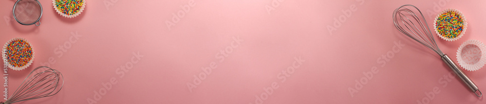 Banner of cupcake liners with colored sugar inside whisk and strainer flat lay top view Confectionery cooking concept with copy space on bright pink paper National cooking day