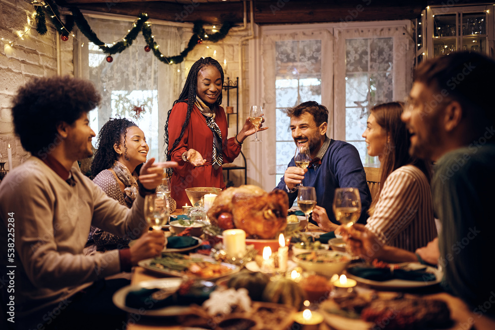 Happy black woman holding toast during Thanksgiving meal with friends at dining table.